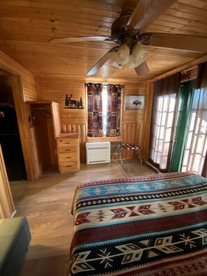 A wooden cabin room with a bed, a ceiling fan, a radiator, and a window with curtains.
