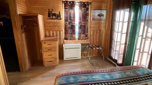Habitació d'una cabana de fusta amb un llit, un ventilador de sostre, un radiador i una finestra amb cortines.