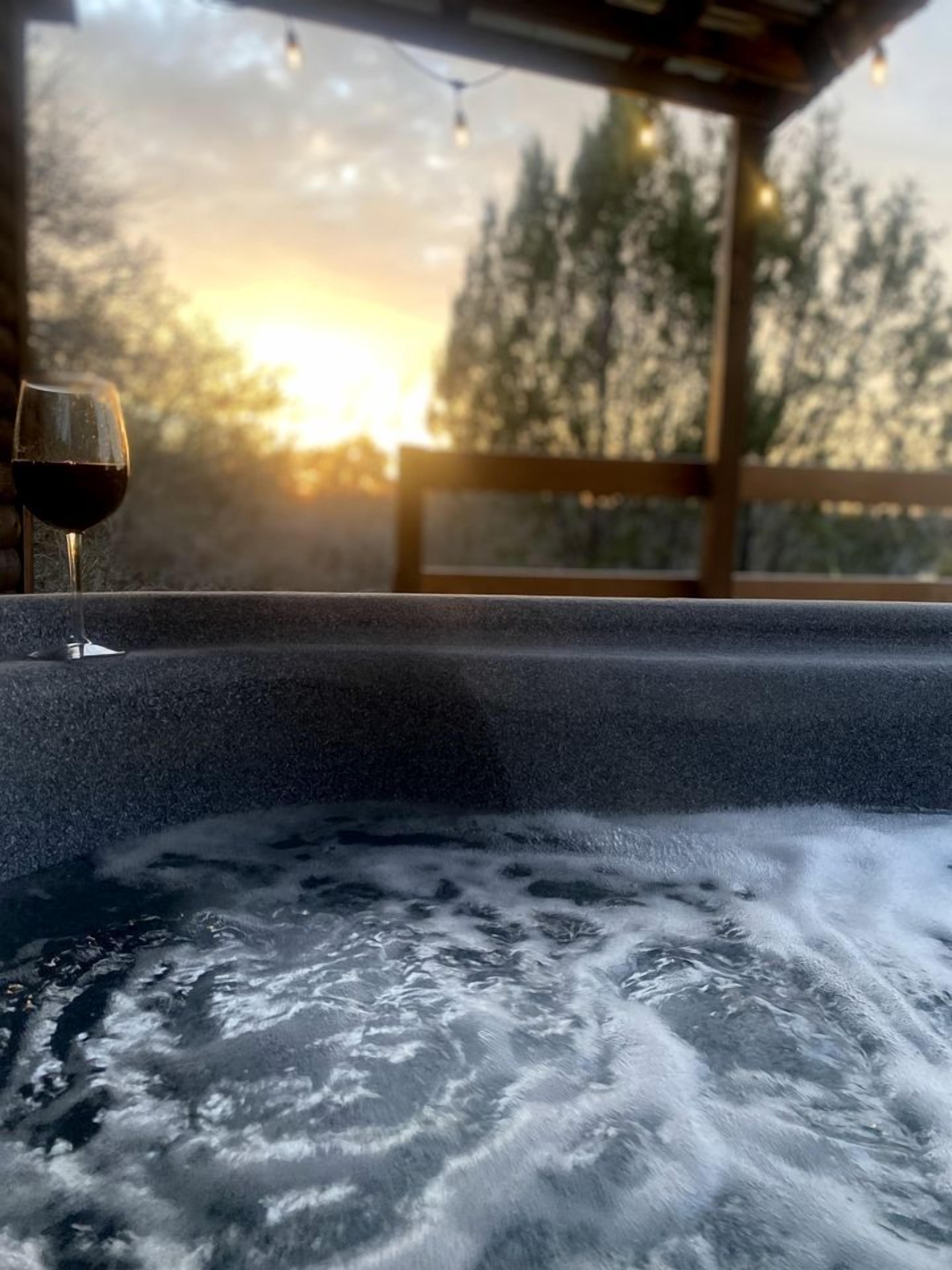 A hot tub with a glass of red wine, surrounded by a wooden deck and trees, with a sunset in the background.