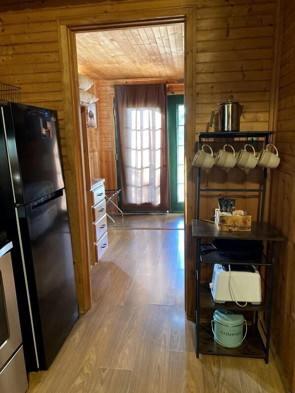 Un interior de madera con una zona de cocina que incluye un refrigerador, una repisa con utensilios de cocina y una ventana con cortinas.