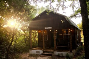 A rustic cabin with a porch, labeled 'YELLASONE,' surrounded by trees and sunlight. - Vacation Cabin Rental with a Private Hot Tub near Fort Worth, Texas (Bluff Dale)