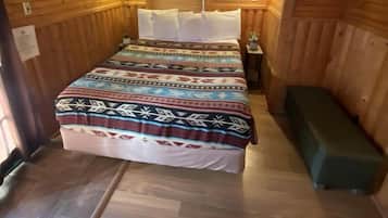 A wooden cabin room with a bed, a ceiling fan, a window with blinds, and a small bedside table with a lamp.