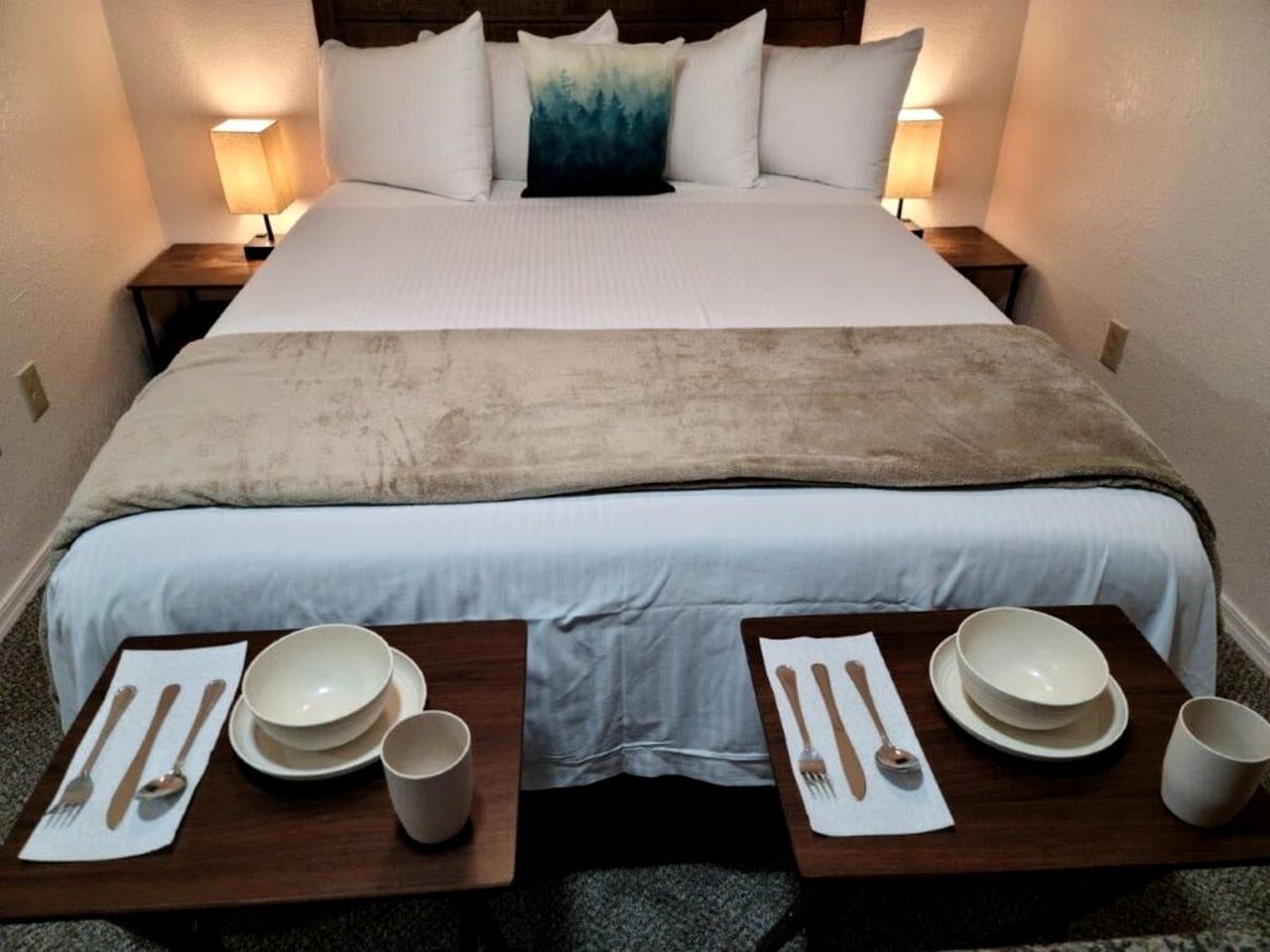 A neatly made bed with a white and blue blanket, two bedside tables with plates, bowls, and cutlery, and two wall-mounted lamps.