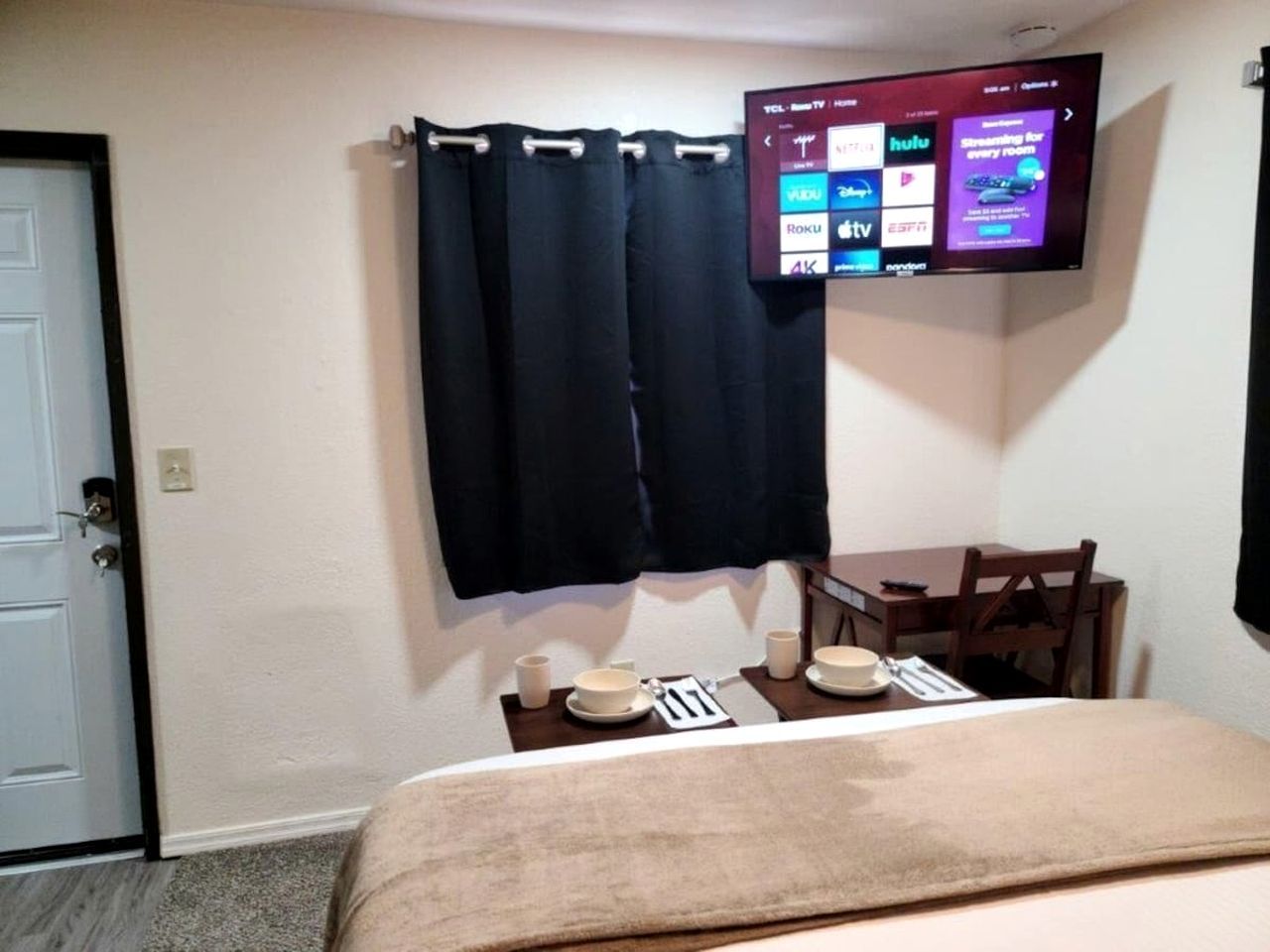 A hotel room with a bed, a small table with a chair, a TV mounted on the wall, and a door.