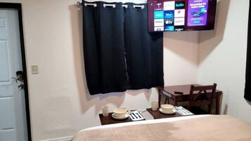 A hotel room with a bed, a small table with a chair, a TV mounted on the wall, and a door.