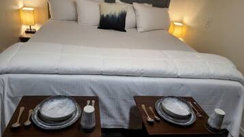 Uma cama bem arrumada com roupa de cama branca, duas mesas de cabeceira com pratos, tigelas e talheres, e duas luminárias fixadas na parede.