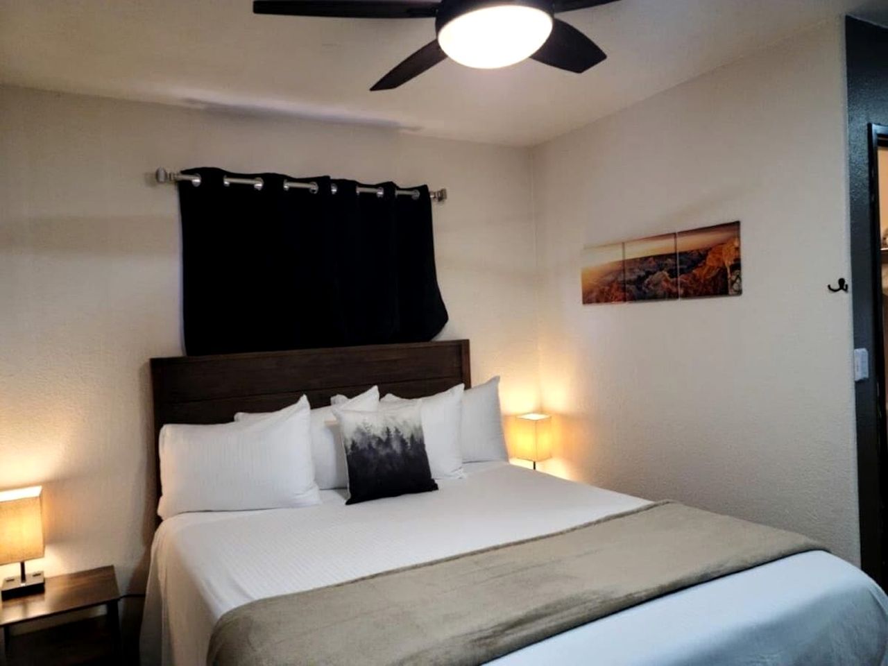 A hotel room with a bed, a ceiling fan, a nightstand, a lamp, and a painting on the wall.