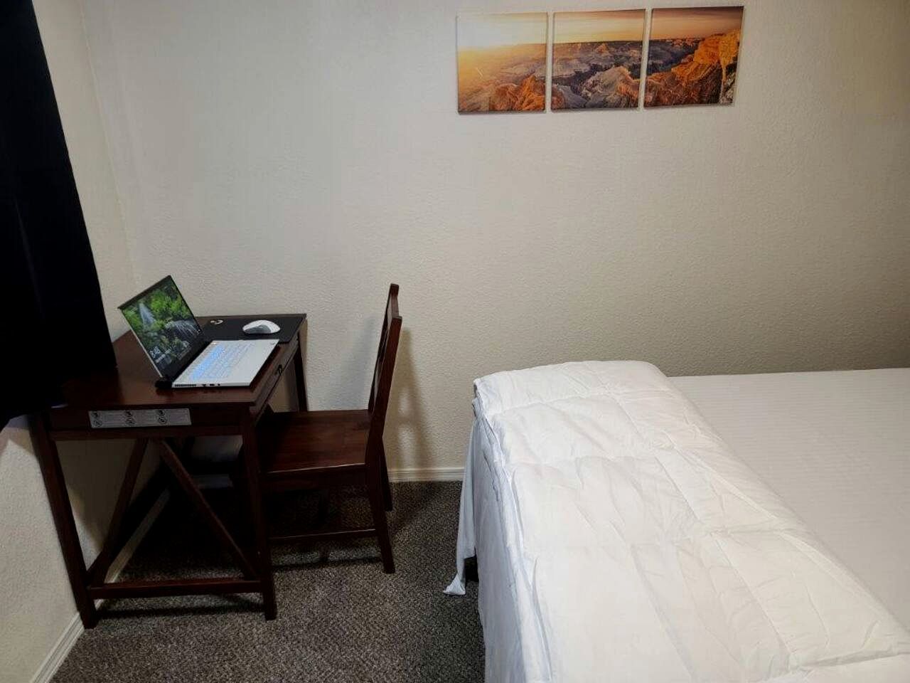 A hotel room with a bed, a desk with a laptop, a chair, and a painting on the wall.