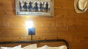A wooden cabin room with a bed, white pillows, a patterned blanket, a mounted painting, and a hat on the wall. - The Renegade in Bluff Dale, TX (Bluff Dale)