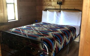 Un llit amb un estovat estampat, estructura de metall i almoines blanques en una habitació amb parets de fusta i una finestra.