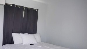 Un lit simple avec un matelas blanc et une tête de lit sombre, une petite table de chevet et un climatiseur fixé au mur.
