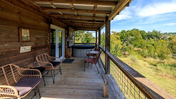 Una cabaña de madera con porche, sillas de mimbre, un jacuzzi y vistas del bosque circundante.