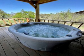 A hot tub on a wooden deck with a clear view of trees and a building in the background. - Beautiful Cabin with Deck and Hot Tub Overlooking Spring Creek in Texas (Fredericksburg)