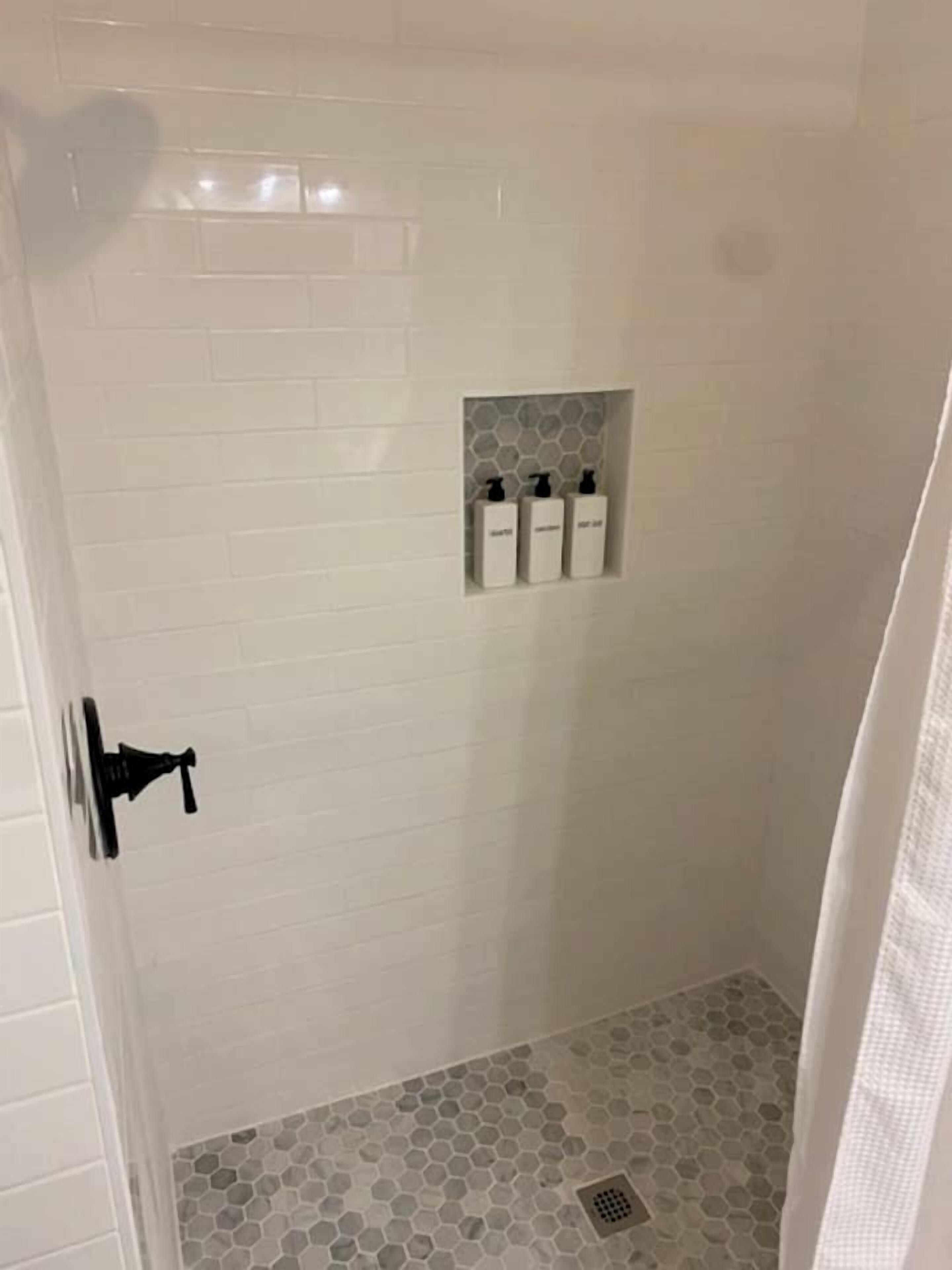 A shower with white tiled walls, a hexagonal floor pattern, and a small shelf with bottles.