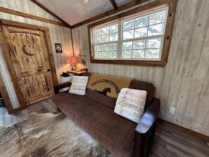 A rustic interior with a wooden door, a window with a view, a sofa with a 'Yellowstone' pillow, and a lamp. - 05) Yellowstone Treehouse (Forestburg)