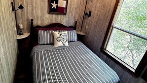 A bedroom with a wooden ceiling, a bed with striped bedding, a Texas flag on the wall, and a window with a view of trees. - Unique Treehouse on Working Farm in Texas, Perfect for Rustic Weekends in Nature (Forestburg)