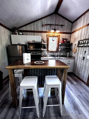 A compact kitchen with wooden cabinets, a stainless steel refrigerator, a sink, and two white stools.