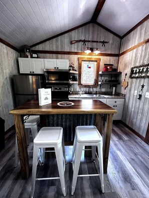 A compact kitchen with wooden cabinets, a stainless steel refrigerator, a sink, and two white stools.