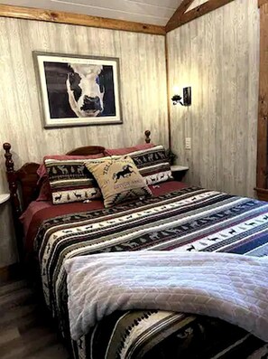 Une chambre à coucher avec une tête de lit en bois, un lit recouvert d’une literie à motifs, un tableau encadré accroché au mur et un luminaire fixé au mur.