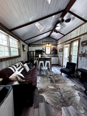 A rustic cabin interior with a wooden ceiling, a sofa, a dining table, and a kitchen area.