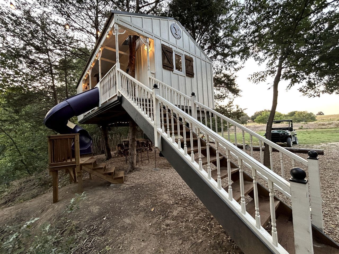 Une cabane avec un escalier menant à une terrasse, un toboggan et un véhicule en arrière-plan.