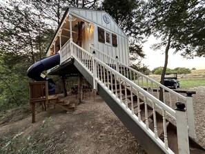 A cabin with a staircase leading to a deck, a slide, and a vehicle in the background. - 05) Yellowstone Treehouse (Forestburg)