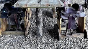 A rustic wooden table under a corrugated metal roof, surrounded by leather saddles and equestrian equipment. - Unique Treehouse on Working Farm in Texas, Perfect for Rustic Weekends in Nature (Forestburg)