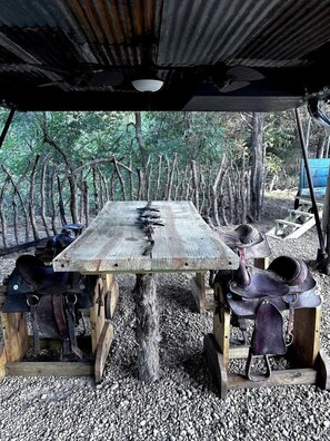 A rustic wooden table under a corrugated metal roof, surrounded by leather saddles and equestrian equipment.