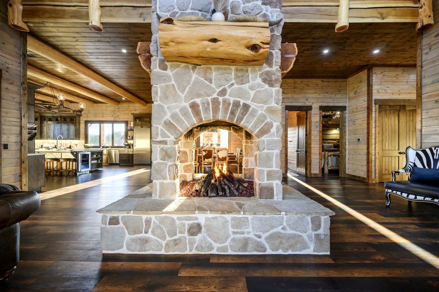 A stone fireplace with a log fire, a wooden ceiling, and a kitchen area in the background.