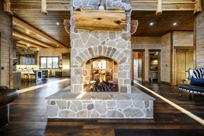A stone fireplace with a log fire, a wooden ceiling, and a kitchen area in the background. - Outstanding Cabin up to 50 People with Bowling, Private Cinema and Fantastic Sightd in Broken Bow, Oklahoma (Broken Bow)