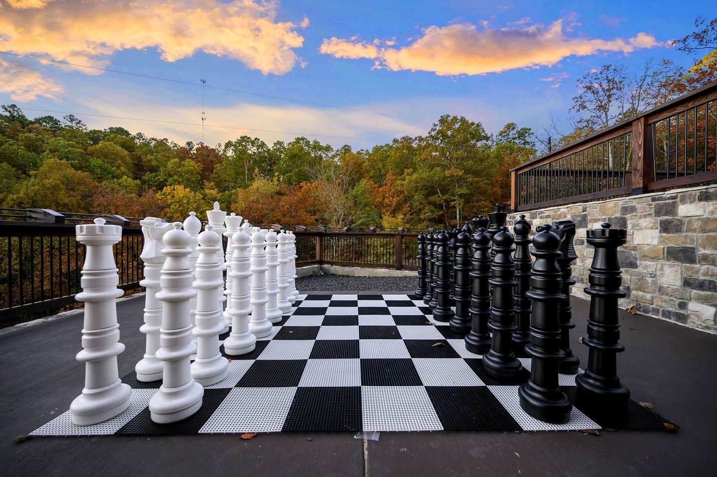 A large outdoor chessboard with oversized chess pieces, surrounded by a stone wall and a wooden fence, set against a backdrop of autumn trees.