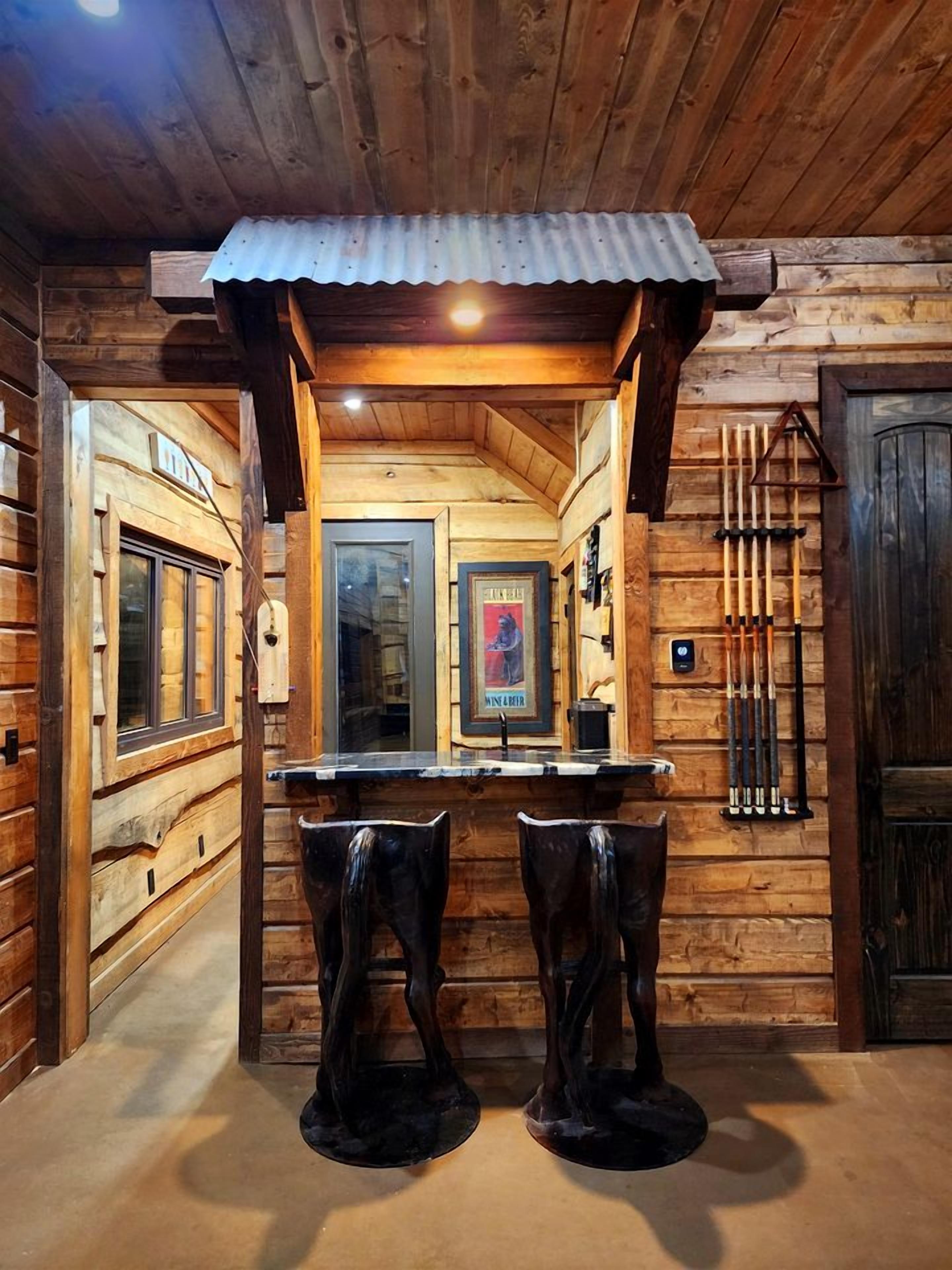 A log cabin interior with a wooden bar area, two stools, a framed picture, and a mounted collection of tools.