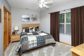 Ein Schlafzimmer mit Bett, Nachttisch, Lampe, Deckenventilator, einem Fenster mit Vorhängen und Wandbildern.