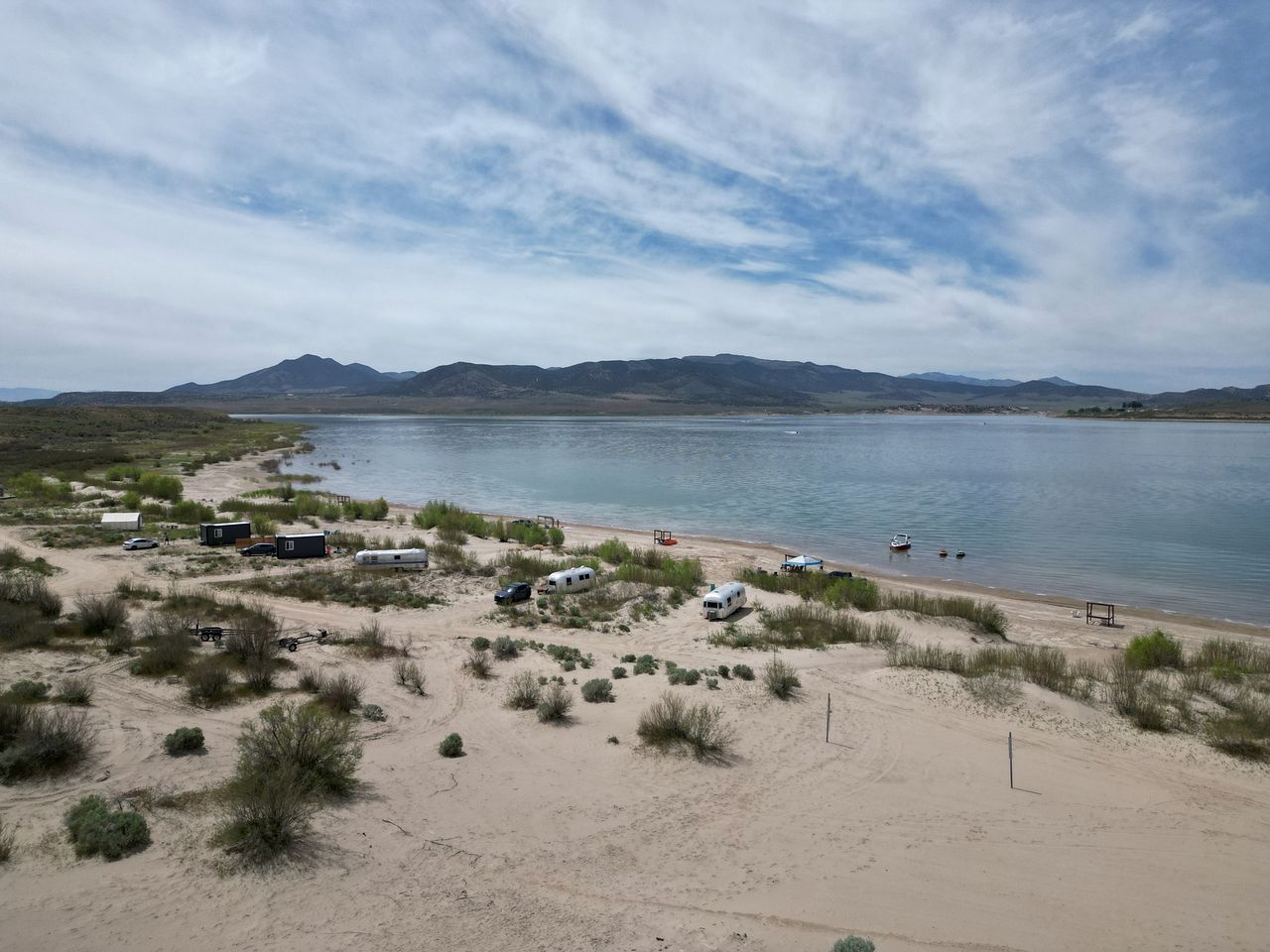 Una playa con remolques de camping y un barco, rodeada de dunas de arena y montañas.
