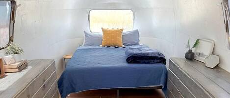 A compact, well-lit interior with a bed, bedside tables, and a small round table with a plant.