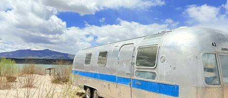 A vintage silver bus parked on a sandy area with a clear sky and mountains in the background.