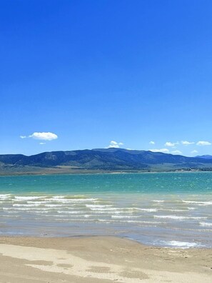 A beach with clear turquoise water, a sandy shore, and distant mountains under a blue sky.