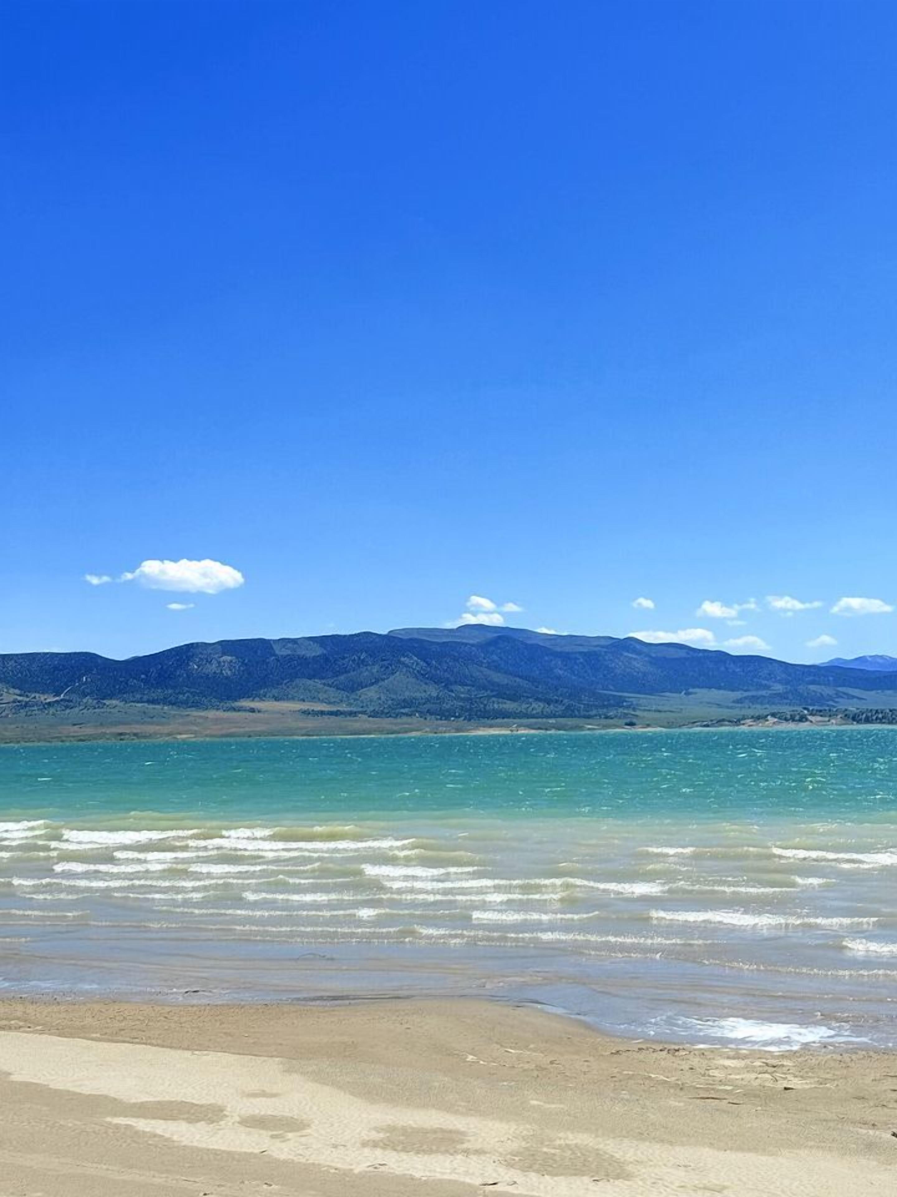 Une plage à l’eau claire, un rivage sablonneux et des montagnes lointaines sous un ciel bleu.