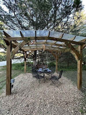 A wooden pergola with a table and chairs underneath, surrounded by gravel.