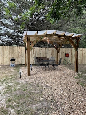 A wooden pergola with a table and chairs underneath, a grill, and a red target stand.