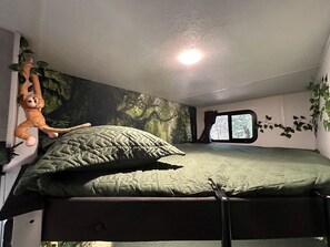 A bunk bed with a green quilted mattress, a hanging plush toy, and a window with a view of trees.