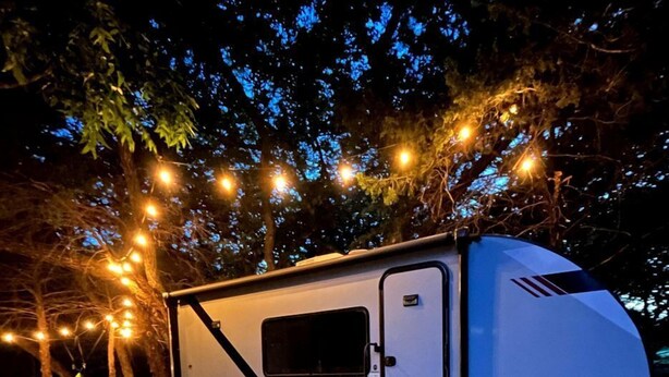A white RV with a 'Platinum' label parked on gravel, surrounded by trees and string lights. - Unique Jungle-Style Glamping RV with 8-Foot Suspended Tree Tent near Creek Trails in Forestburg, Texas (Forestburg)