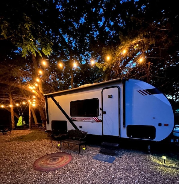 A white RV with a 'Platinum' label parked on gravel, surrounded by trees and string lights.