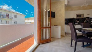 Uma varanda com vista para edifícios, uma porta de correr de vidro, uma área de cozinha com armários e micro-ondas, e uma mesa de jantar com cadeiras.