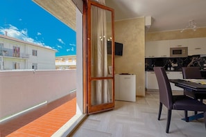 Un balcón con vistas a edificios, una puerta corrediza de vidrio, una zona de cocina con armarios y un microondas, y una mesa de comedor con sillas.
