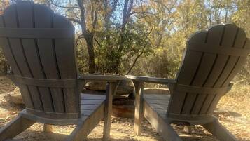 Two Adirondack chairs facing each other with a table in between, set in a natural outdoor area with trees and a clear sky.