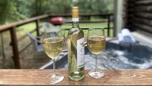 Une bouteille de vin et deux verres sur une table en bois, avec vue sur une terrasse en bois et sur la verdure.