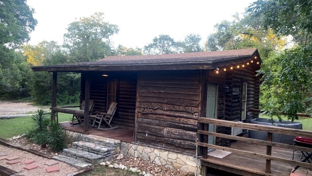 Une cabane rustique en rondins de bois, dotée d’un porche, de sièges extérieurs et d’un foyer.