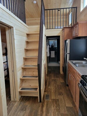 Una escalera de madera estrecha que sube a un segundo piso, una cocina con armarios de madera y un refrigerador negro, y una puerta de baño visible.