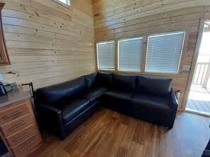 A room with a black leather sectional sofa, wooden flooring, a wooden cabinet, and two windows with blinds. - Quiet Desert Escape in a Stylish Tiny Home with Starry Skies in Williams, Arizona (Williams)